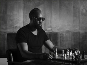 "Man wearing sunglasses and a dark t-shirt sits at a table concentrating on a chess game in a dimly lit, modern room with abstract patterned walls. The scene is in black and white, evoking a thoughtful, serious mood."