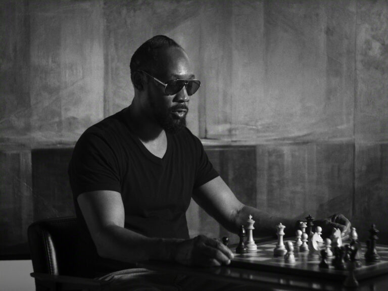 "Man wearing sunglasses and a dark t-shirt sits at a table concentrating on a chess game in a dimly lit, modern room with abstract patterned walls. The scene is in black and white, evoking a thoughtful, serious mood."