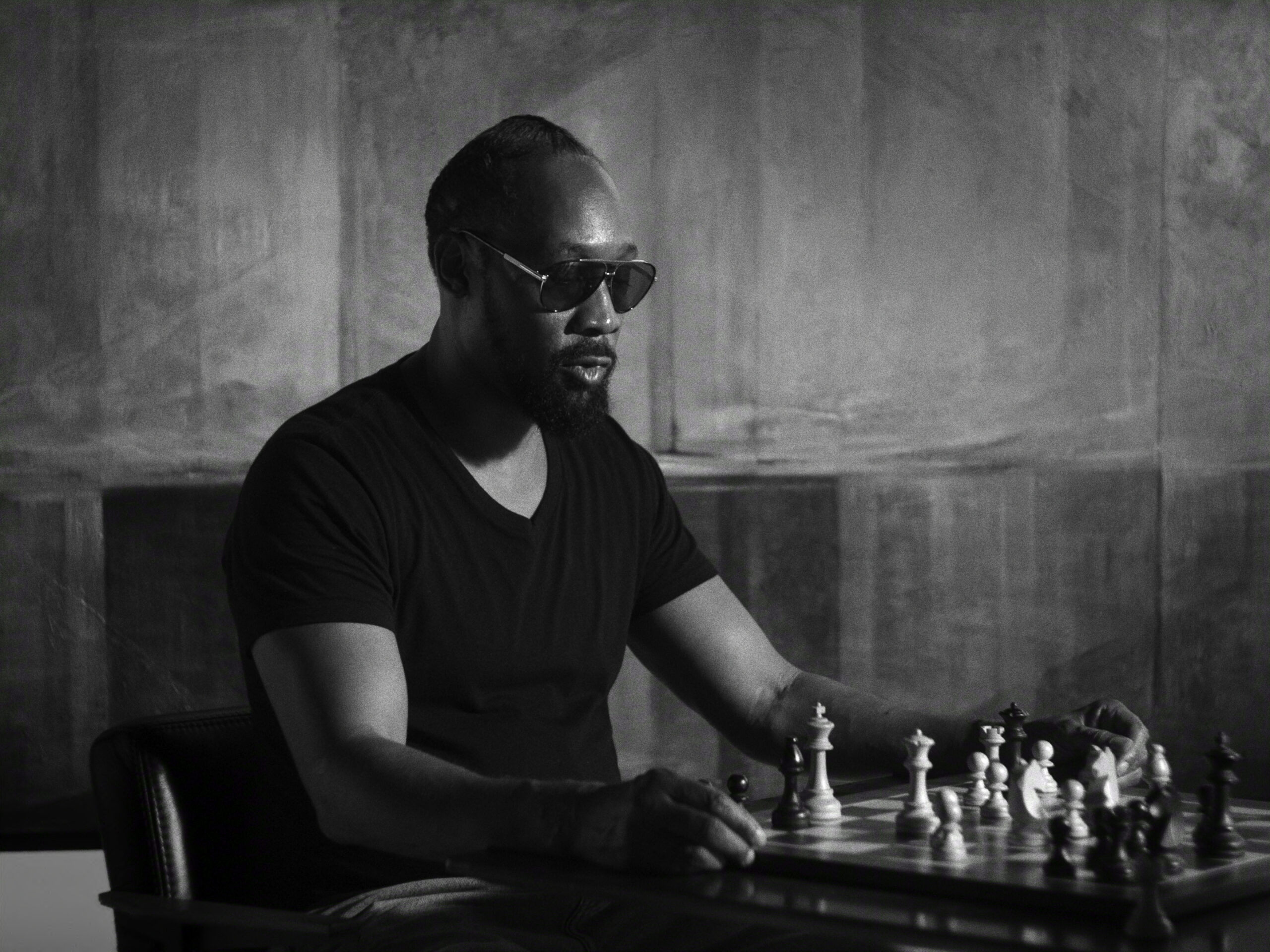 "Man wearing sunglasses and a dark t-shirt sits at a table concentrating on a chess game in a dimly lit, modern room with abstract patterned walls. The scene is in black and white, evoking a thoughtful, serious mood."